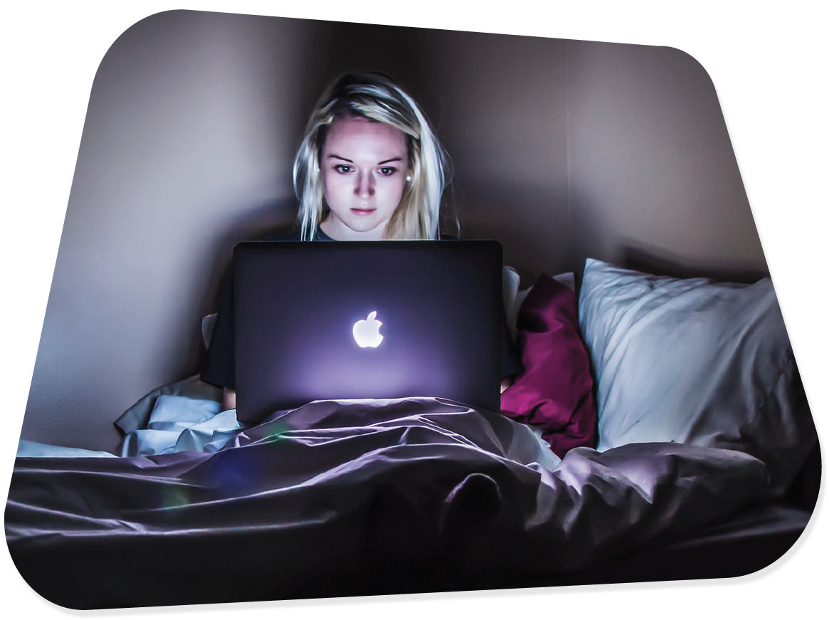A picture of a teenage girl using her laptop from bed, in a dark room with the laptop illuminating her face.