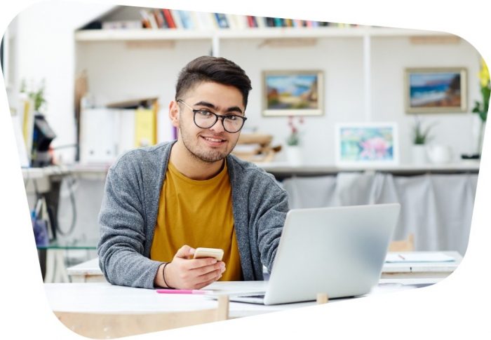 A young man wearing glasses is sat at a desk, holding a phone in one hand and using a laptop with the other.